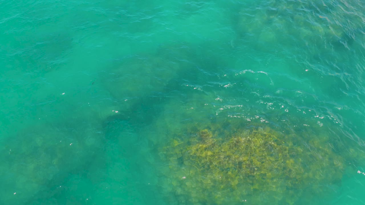 At 0:02, clear turquoise water in Laguna Beach, Baja California Sur shows a submerged rock patch lower in frame and brighter ripple highlights near the surface, with no visible people.