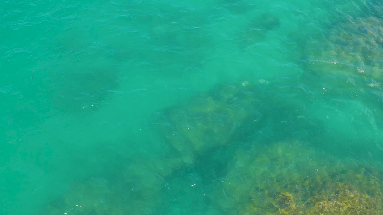 Overhead view of clear turquoise sea at Laguna Beach, Baja California Sur, with underwater rock patches concentrated toward the lower right and no visible people.