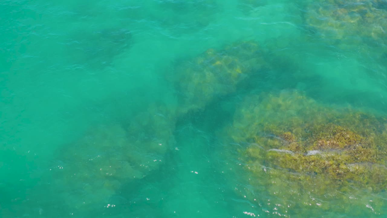 Aerial frame over clear green lagoon water in Laguna Beach, Baja California Sur, Mexico, with a larger rock patch along the right side and faint ripples across the surface, no visible people.