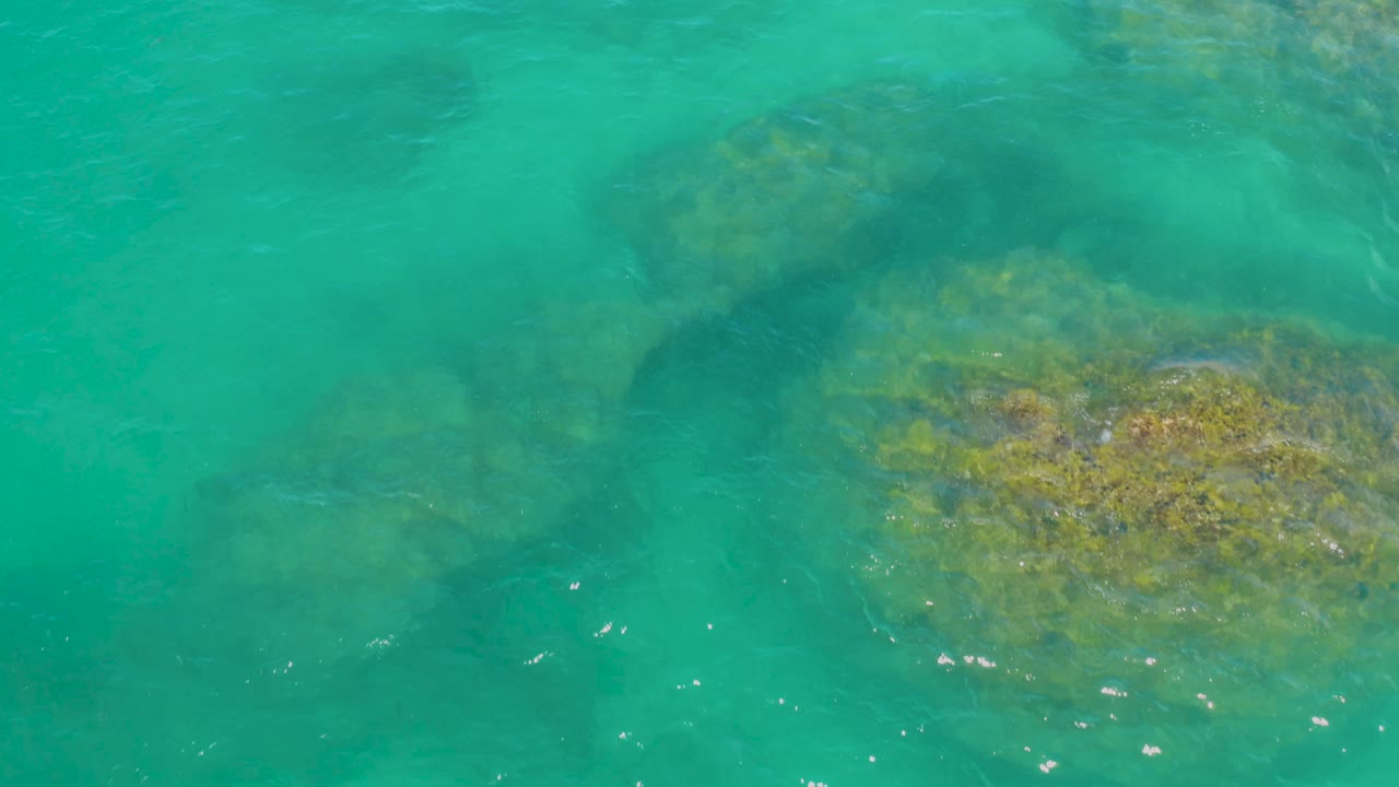 Top-down lagoon view in Laguna Beach, Baja California Sur, Mexico, showing turquoise-green water, a broad underwater rock area on the right, and open water on the left with no visible people.