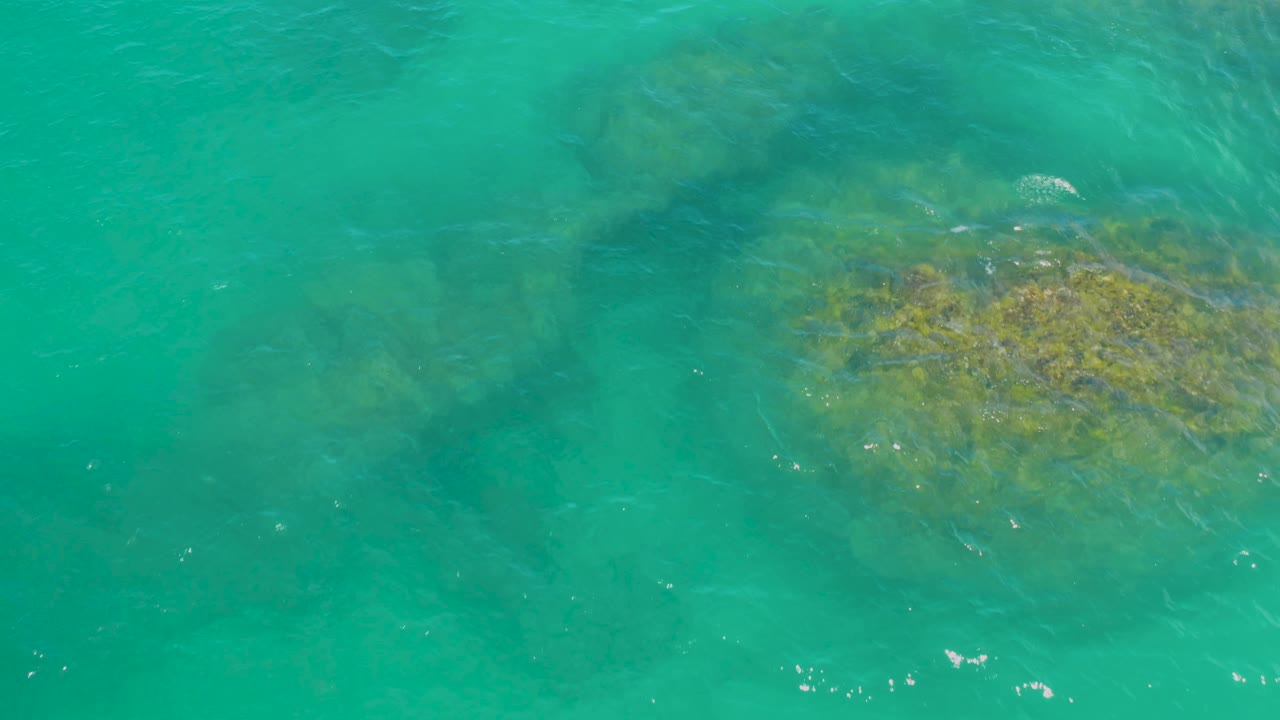 Aerial view of green lagoon water in Laguna Beach, Baja California Sur, Mexico, with a submerged rock patch shifted toward the lower right and small wave lines, no visible people.