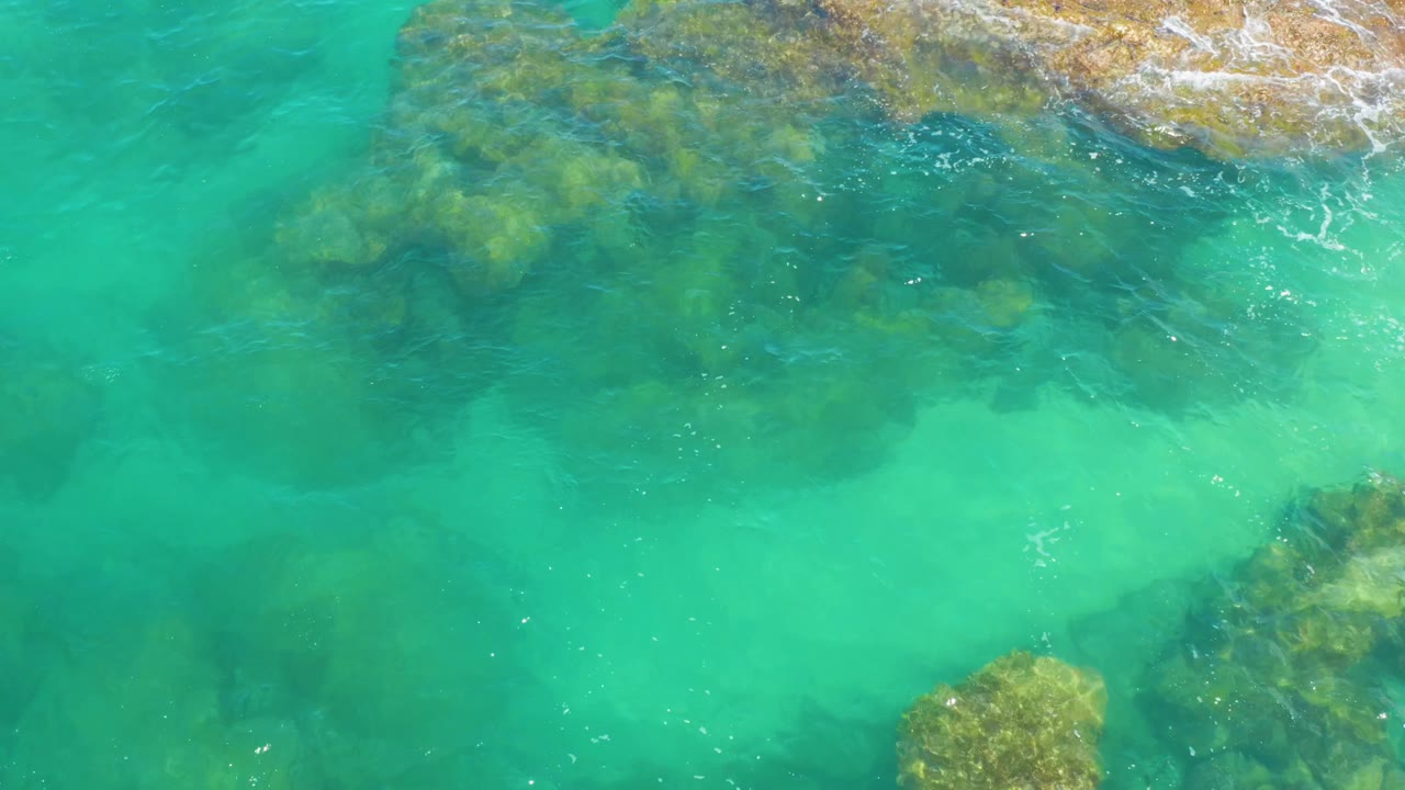 At 0:01, Laguna Beach, Baja California Sur appears from above with green-blue water covering darker rock patches, a tan rocky edge at the top, light surf, and no visible people.