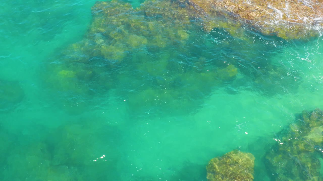 At 0:02, the overhead view of Laguna Beach, Baja California Sur shows aqua water, shadowed rock shapes below the surface, a rough rock edge near the top, small foam streaks, and no visible people.