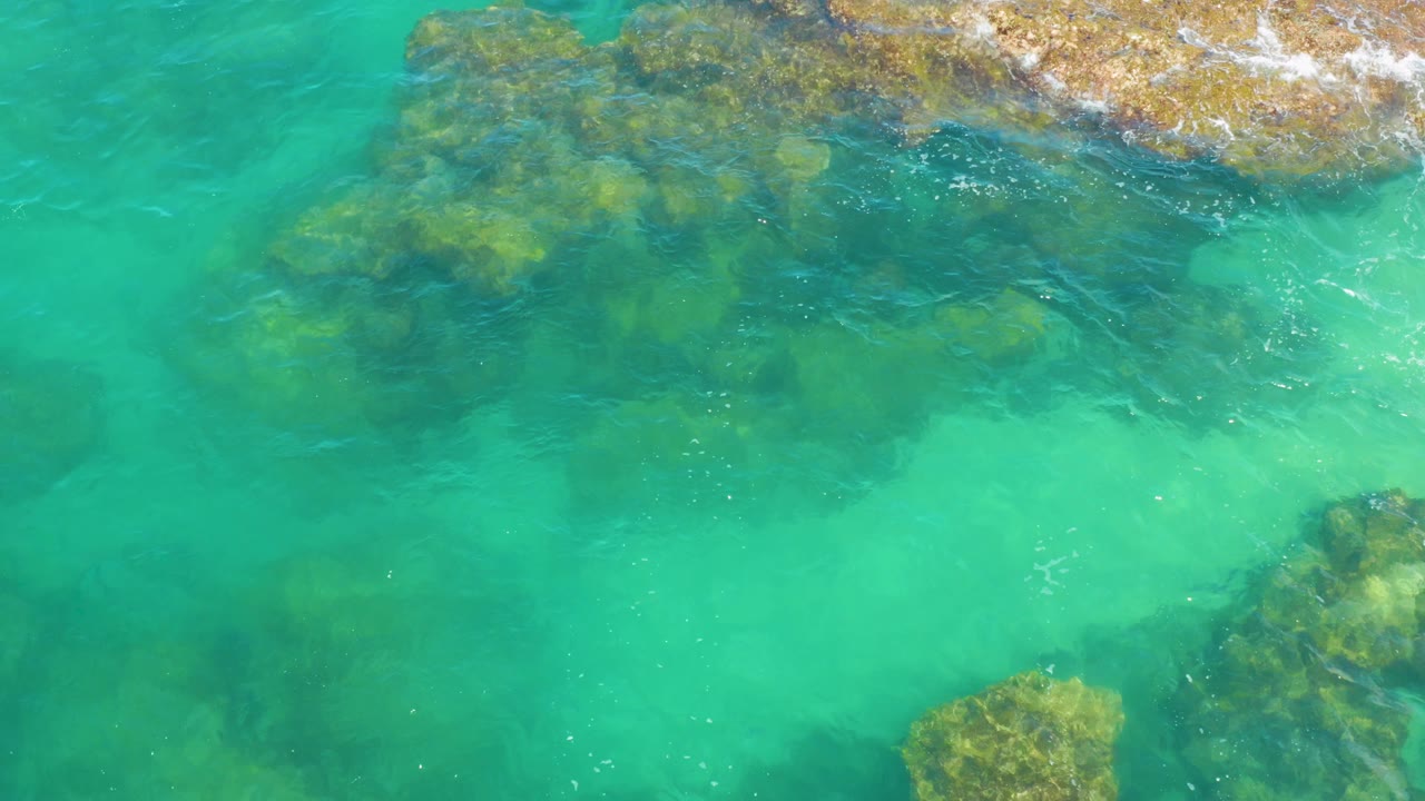 At 0:03, Laguna Beach, Baja California Sur is shown from above with a broad submerged rock formation crossing the upper frame, clear aqua shallows below it, and no visible people.