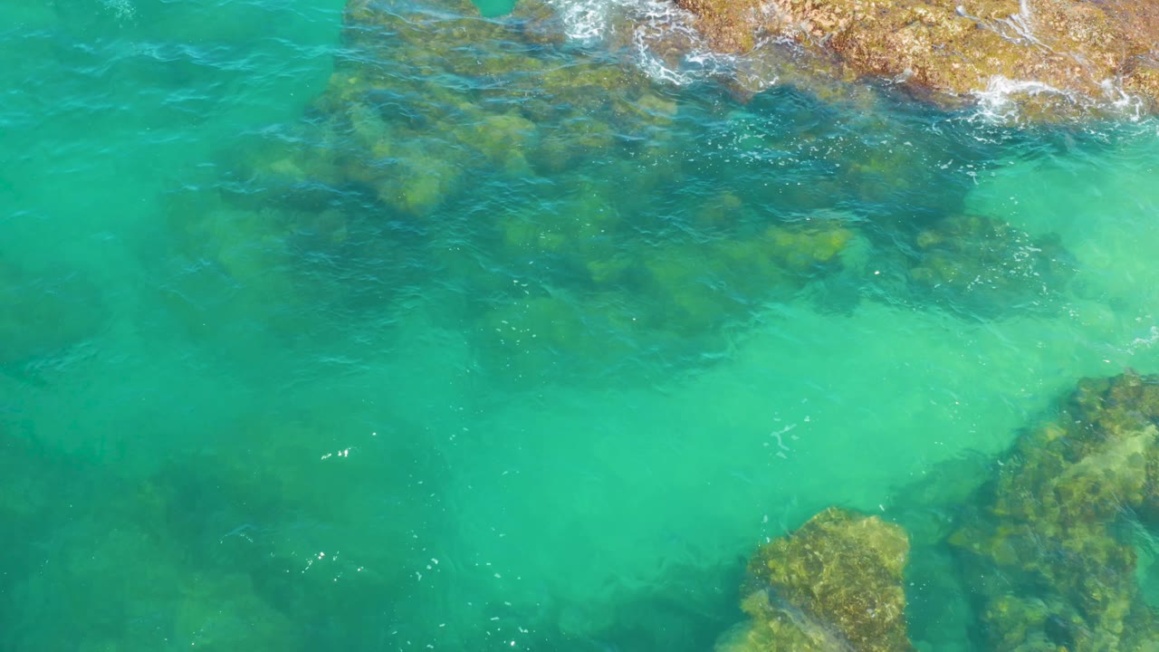 Overhead frame from Laguna Beach, Baja California Sur, with rocky patches near the top and lower right separated by a clear turquoise channel, with no visible people.