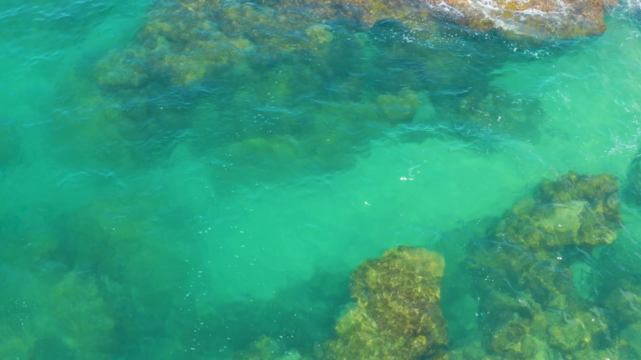Top-down view of blue-green coastal water with submerged rock forms and tiny white foam marks near the upper edge, no visible people.