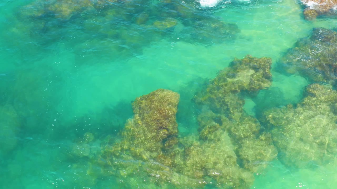 Clear green-blue water in Laguna Beach, Baja California Sur, Mexico, reveals a central cluster of submerged rocks beneath light surface ripples, with no visible people.