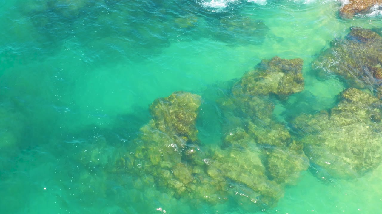 Aerial-looking overhead frame of shallow turquoise water in Laguna Beach, Baja California Sur, Mexico, with submerged rocks drifting lower in the composition and no visible people.