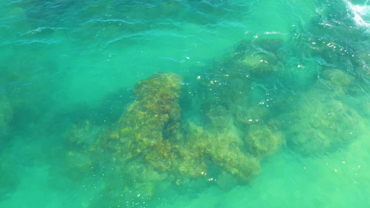 Aerial frame showing green-blue ocean water with a darker submerged reef mass near center and pale sand visible around it at Laguna Beach, with no visible people.
