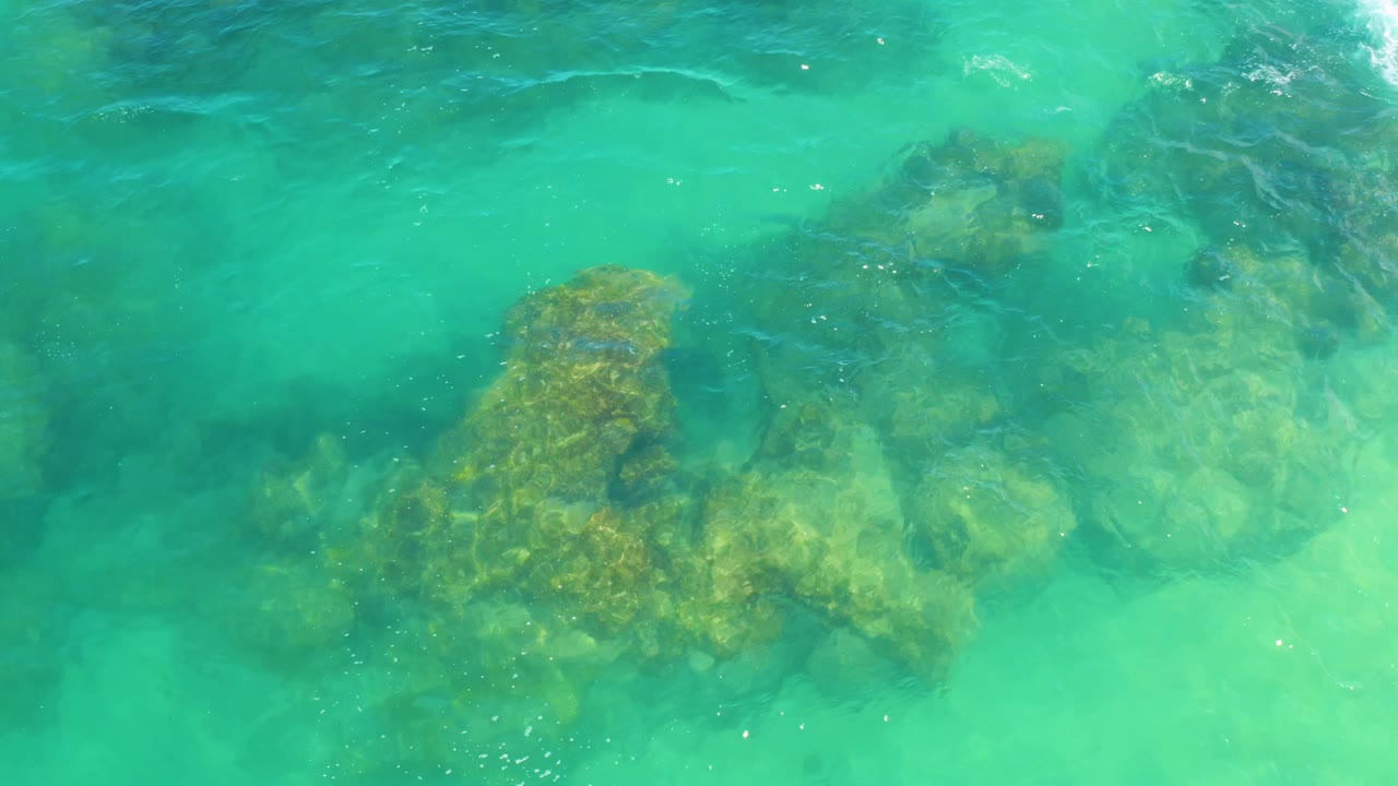 Bright shallow ocean water reveals reef formations spread across the frame at Laguna Beach, Baja California Sur, with a white ripple near the top and no visible people.