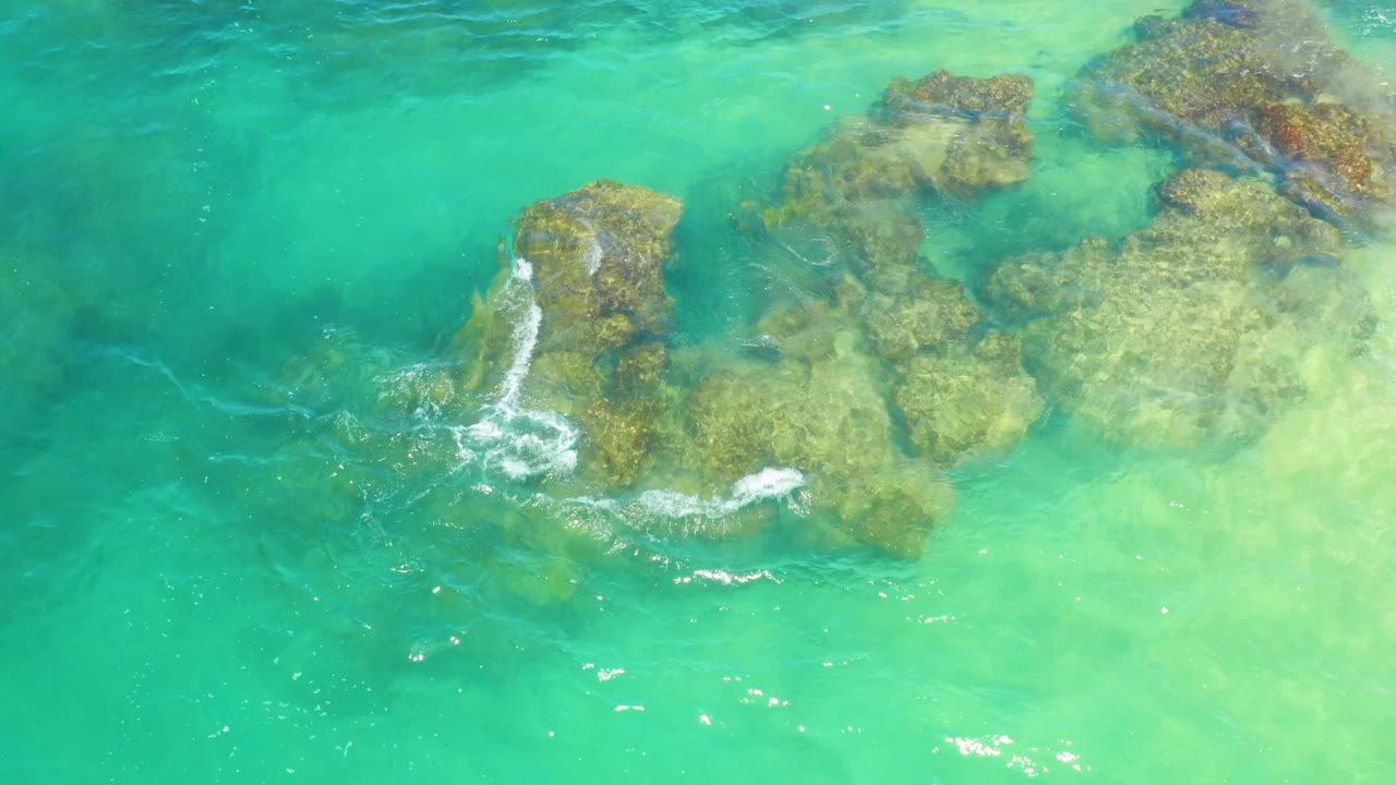 White surf crosses the upper part of a submerged rock formation in Laguna Beach, Baja California Sur, with green water surrounding the rocks and no visible people.
