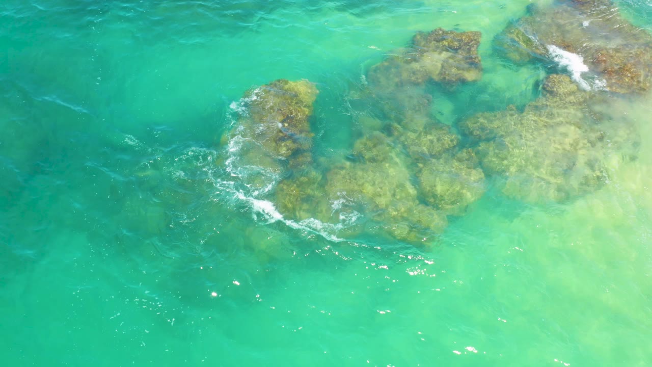 Foam breaks over the left side of the underwater rocks in Laguna Beach, Baja California Sur, with the surrounding water shifting from teal to pale green and no visible people.