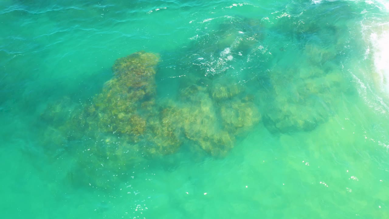 Aerial frame of shallow turquoise water, submerged rocks, and white foam entering from the right at Laguna Beach, Baja California Sur, with no visible people.