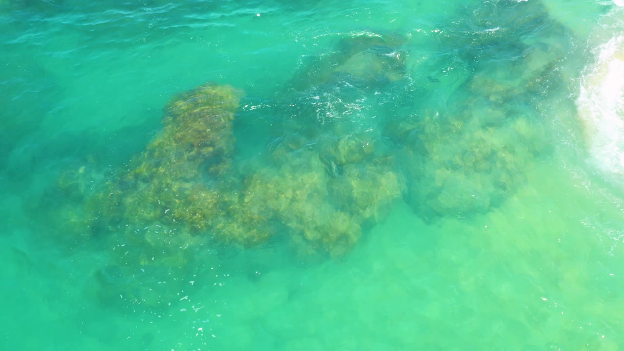 Submerged coastal rocks sit beneath milky turquoise water while white wash reaches the right edge at Laguna Beach, Baja California Sur, with no visible people.