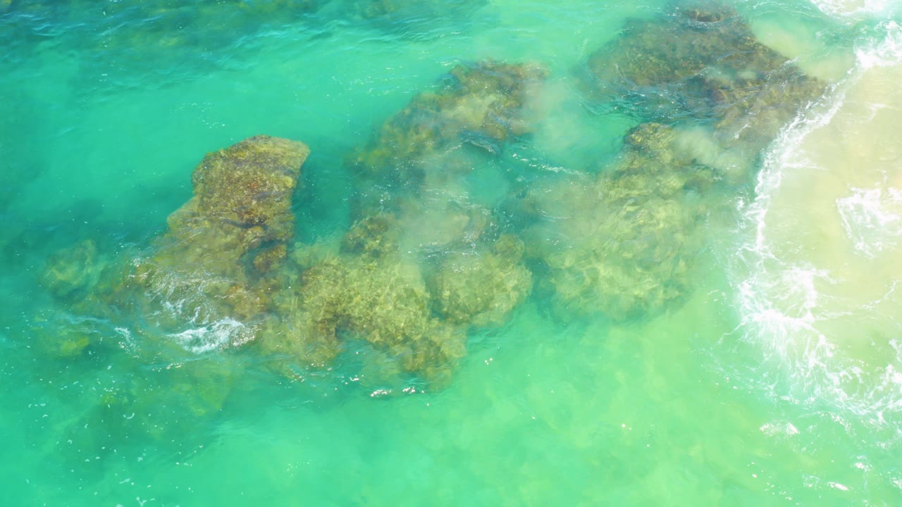 Overhead view in Laguna Beach, Baja California Sur, with clear turquoise water over large submerged rocks, pale sand at right, and no visible people.