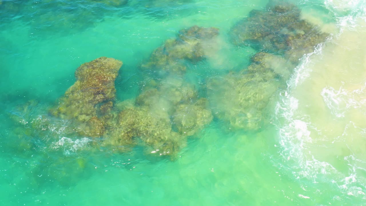 Laguna Beach, Baja California Sur appears from above with a rock cluster under shallow turquoise surf, brighter foam on the right, and no visible people.