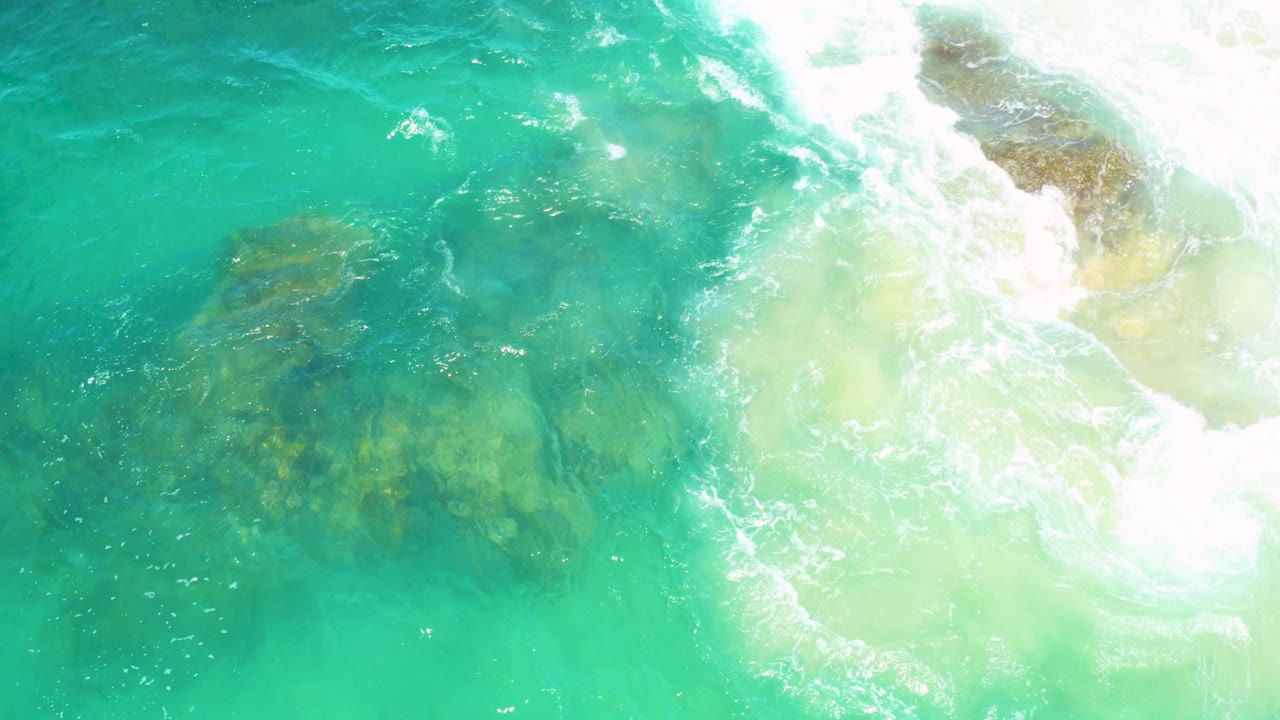 Overhead view of turquoise water washing over submerged rocks in Laguna Beach, Baja California Sur, with bright foam on the right and no visible people.