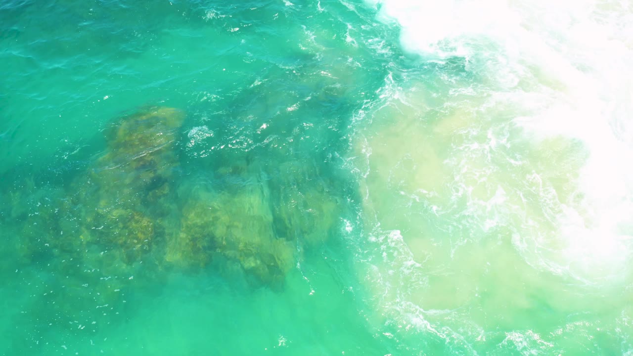 A brighter wash of white foam crosses the upper right while submerged rocks remain visible through turquoise water in Laguna Beach, Baja California Sur, with no visible people.