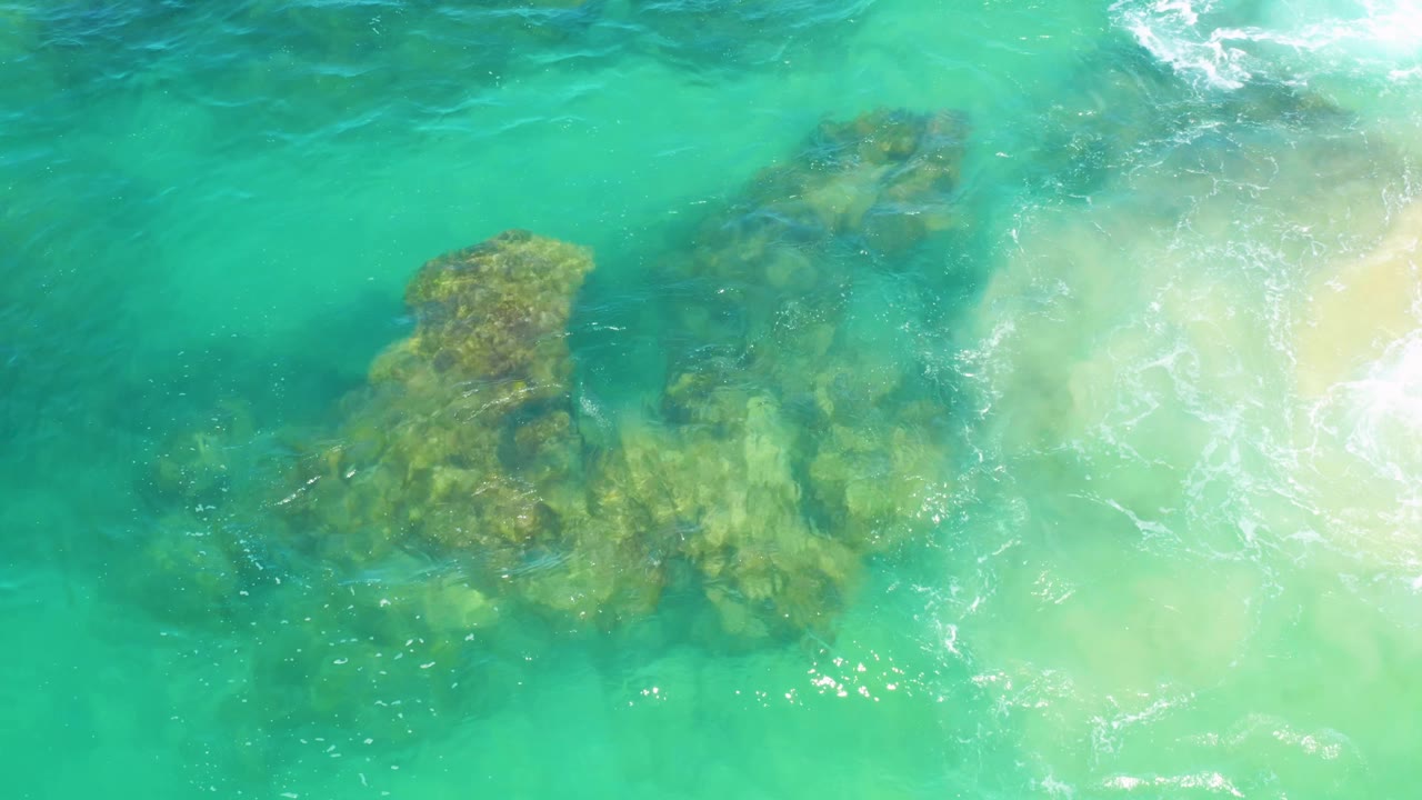 Clear water at Laguna Beach, Baja California Sur reveals textured rock below the surface, with white foam beginning to spread along the right edge and no visible people.