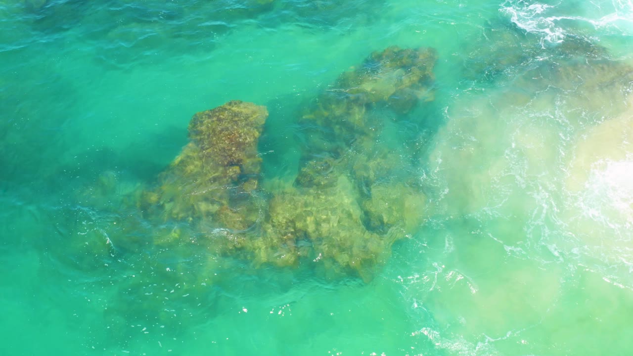 A rocky outcrop sits under bright turquoise water at Laguna Beach, Baja California Sur, while surf foam and sandy shallows fill the right side with no visible people.