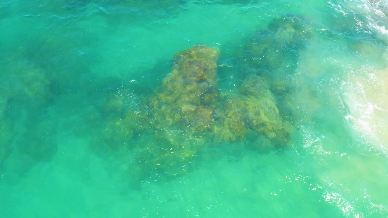 Overhead view of turquoise water off Laguna Beach, Baja California Sur, with a yellow-brown submerged reef centered under the surface, white foam along the right edge, and no visible people.