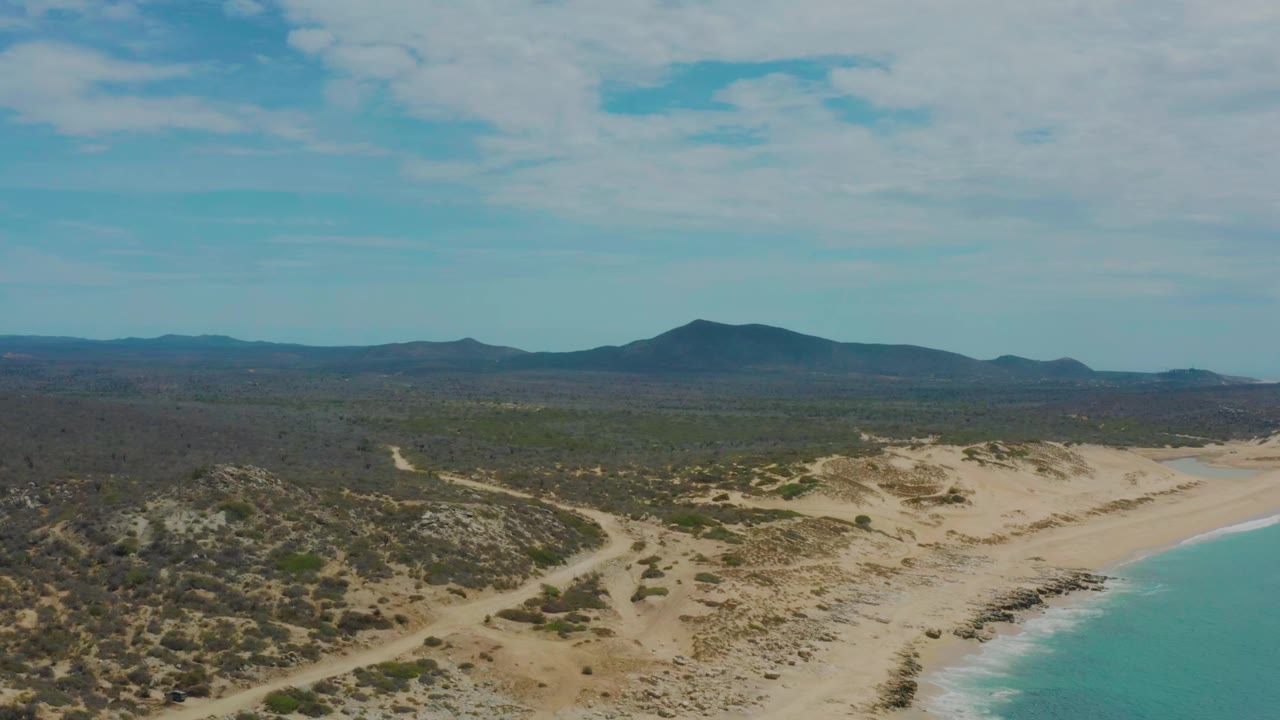 Aerial view over Laguna Beach in Baja California Sur, with scrubby dunes, a curving turquoise shoreline, distant mountains, scattered clouds, and no visible people.