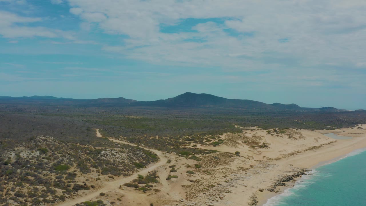 Aerial frame over Laguna Beach, Baja California Sur, showing sandy dunes beside blue-green surf, scrubland inland, distant low mountains, and no visible people.