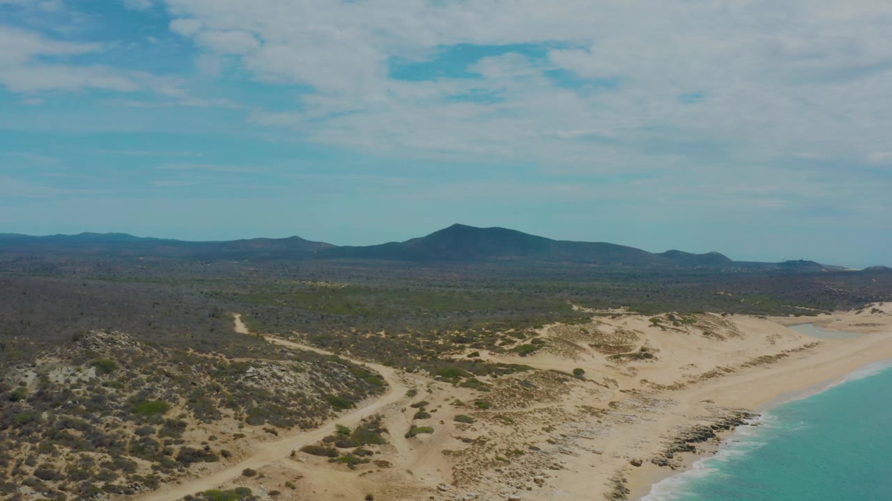 Wide aerial coastline with pale sand, low dunes, surf, scrubland, and mountains.