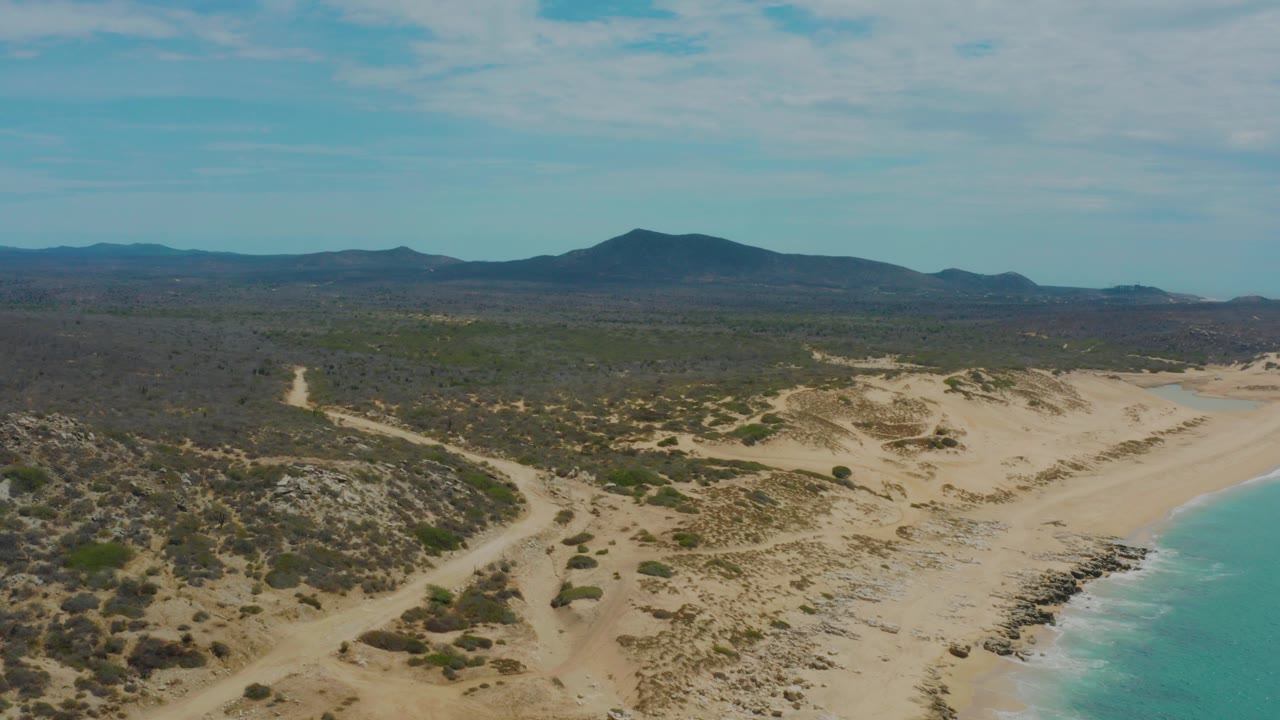 Opening aerial frame over Laguna Beach, Baja California Sur, with turquoise ocean at the right, pale sand dunes below, scrubland and dirt tracks inland, distant mountains, and no visible people.