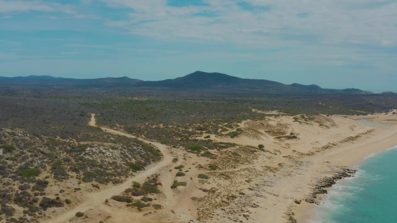 High coastal view with beach curving beside blue water and scrubland leading to mountains.