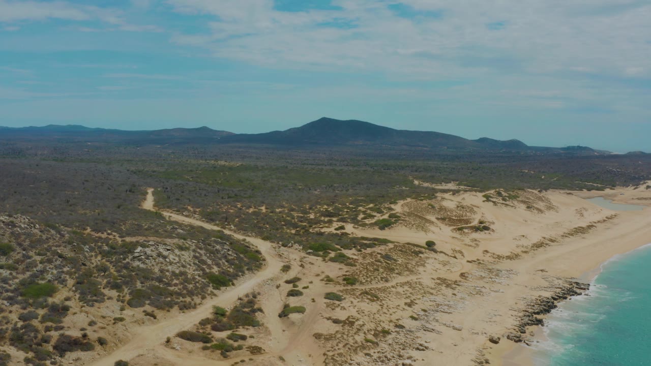 Frame 0:02 shows Laguna Beach, Baja California Sur from above, with a rocky strip at the waterline, pale sand across the center, low brush inland, mountain silhouettes on the horizon, and no visible people.