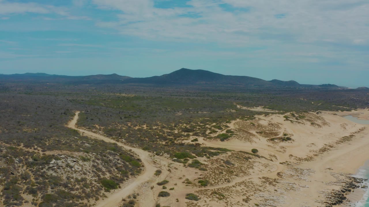 Opening aerial frame over Laguna Beach, Baja California Sur, showing pale sand paths, low green scrub, distant dark mountains, thin clouds, and no visible people.