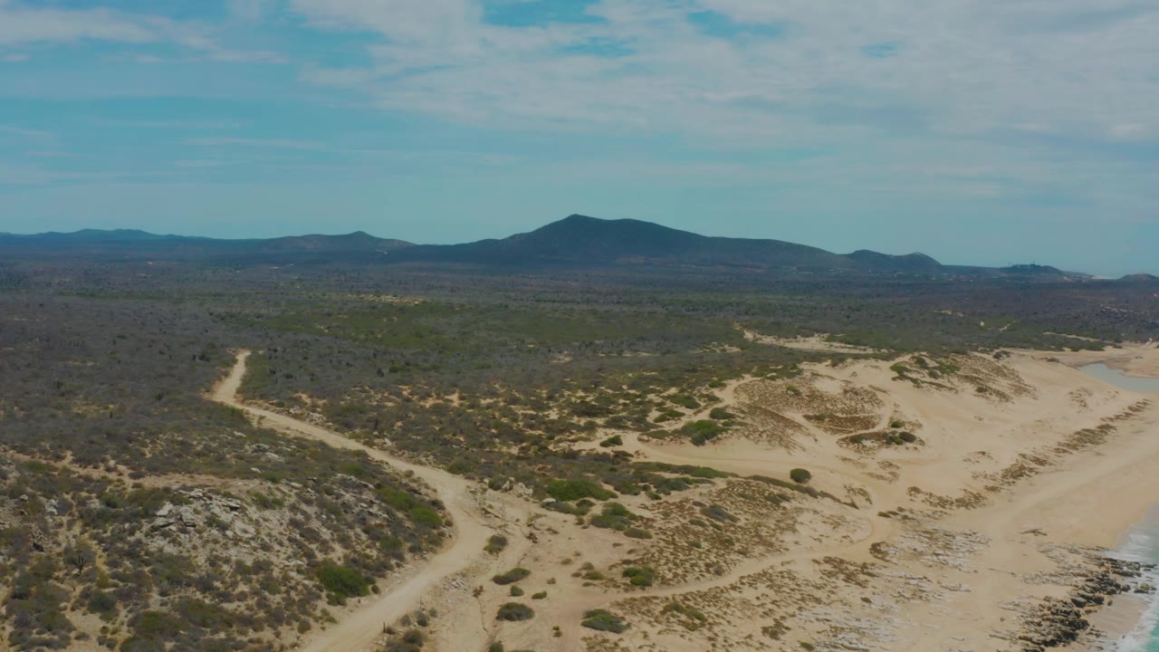 Aerial view in Laguna Beach, Baja California Sur, with a pale dirt track curving through scrub-covered desert toward low mountains, no visible people.