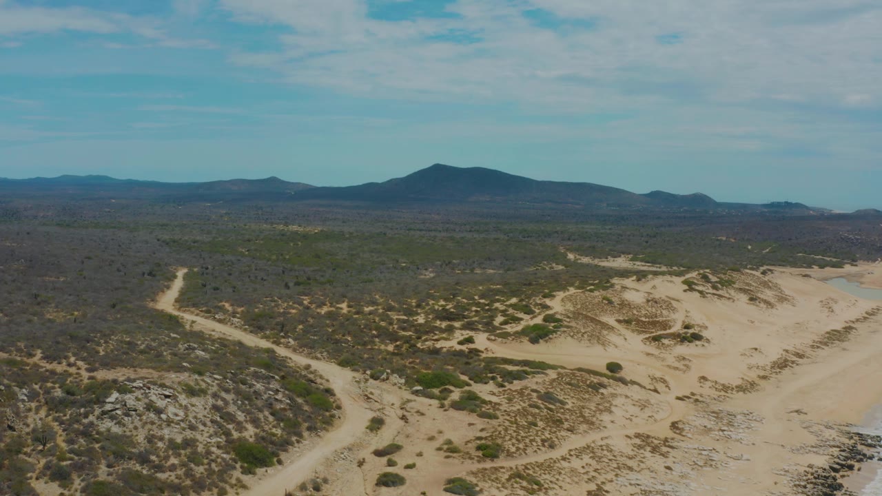 Laguna Beach aerial frame showing broad sandy washes, scattered green plants, darker desert plain, distant mountains, light cloud cover, and no visible people.