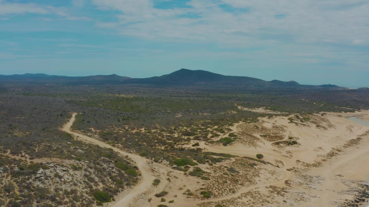 Mid-clip aerial view over Laguna Beach, Baja California Sur, with sandy foreground ridges, scattered brush, a dark mountain ridge, blue sky, and no visible people.