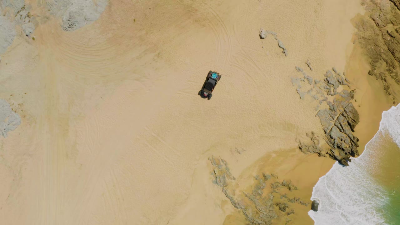 Aerial frame of a dark car crossing open sand, with scattered rocks to the right and a narrow band of foamy surf at Laguna Beach, Baja California Sur, with no visible people.