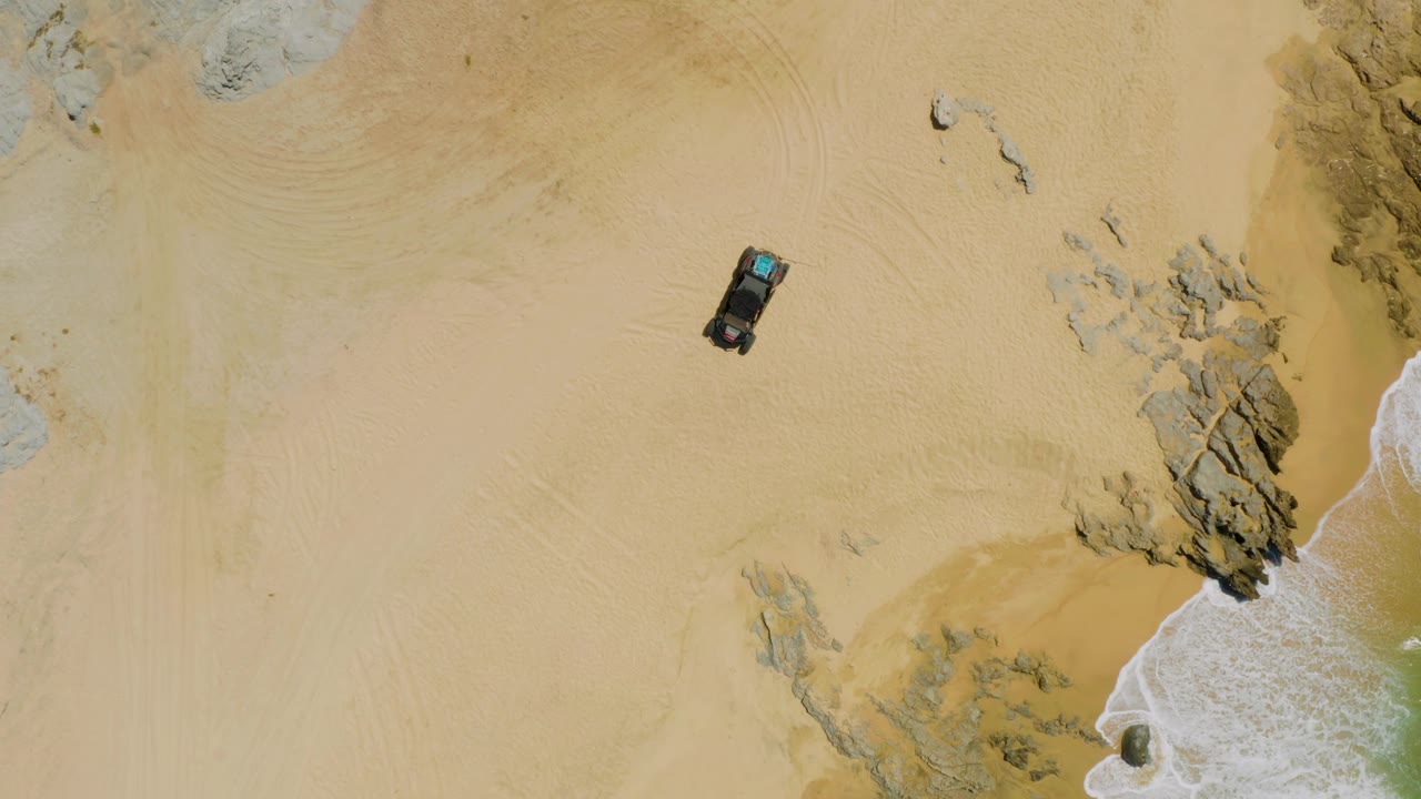 Overhead beach view at Laguna Beach, Baja California Sur, showing the car left of rock formations, pale sand marked by circular tire tracks, and surf along the lower right edge, with no visible people.