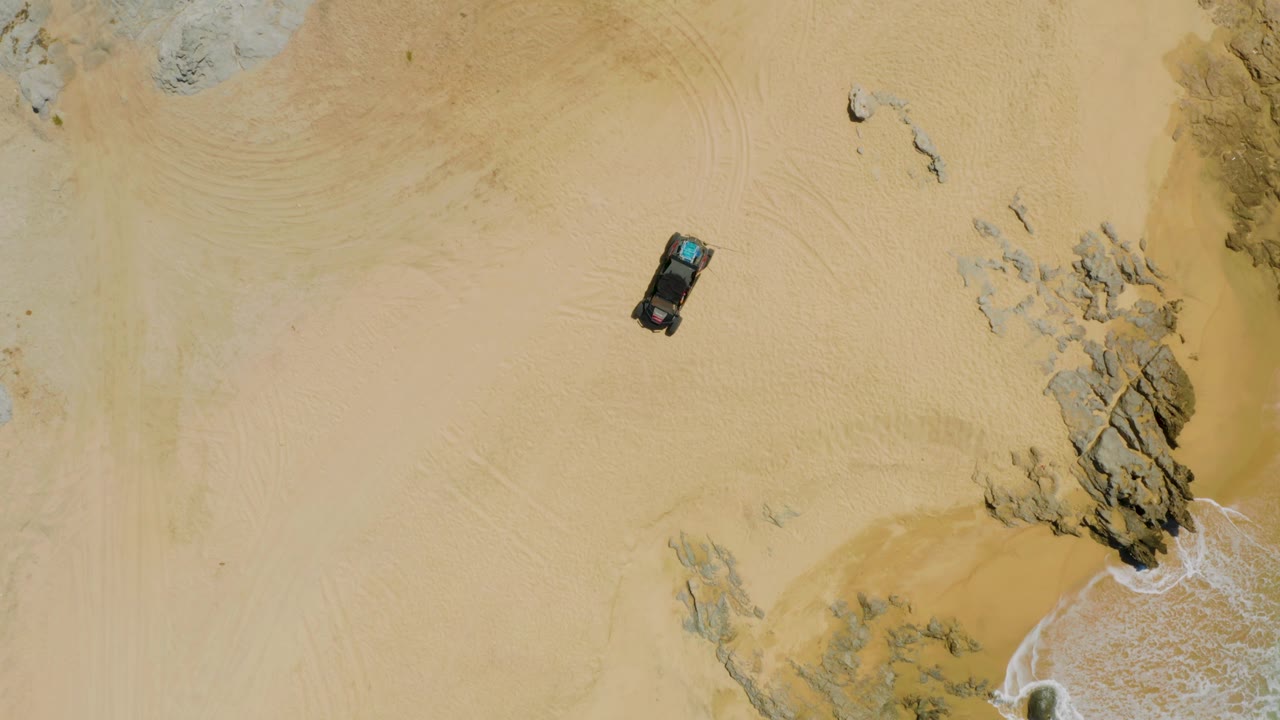 Top-down frame of the car on light sand near clustered rocks, with curved tire marks arcing across the beach at Laguna Beach, Baja California Sur, and no visible people.