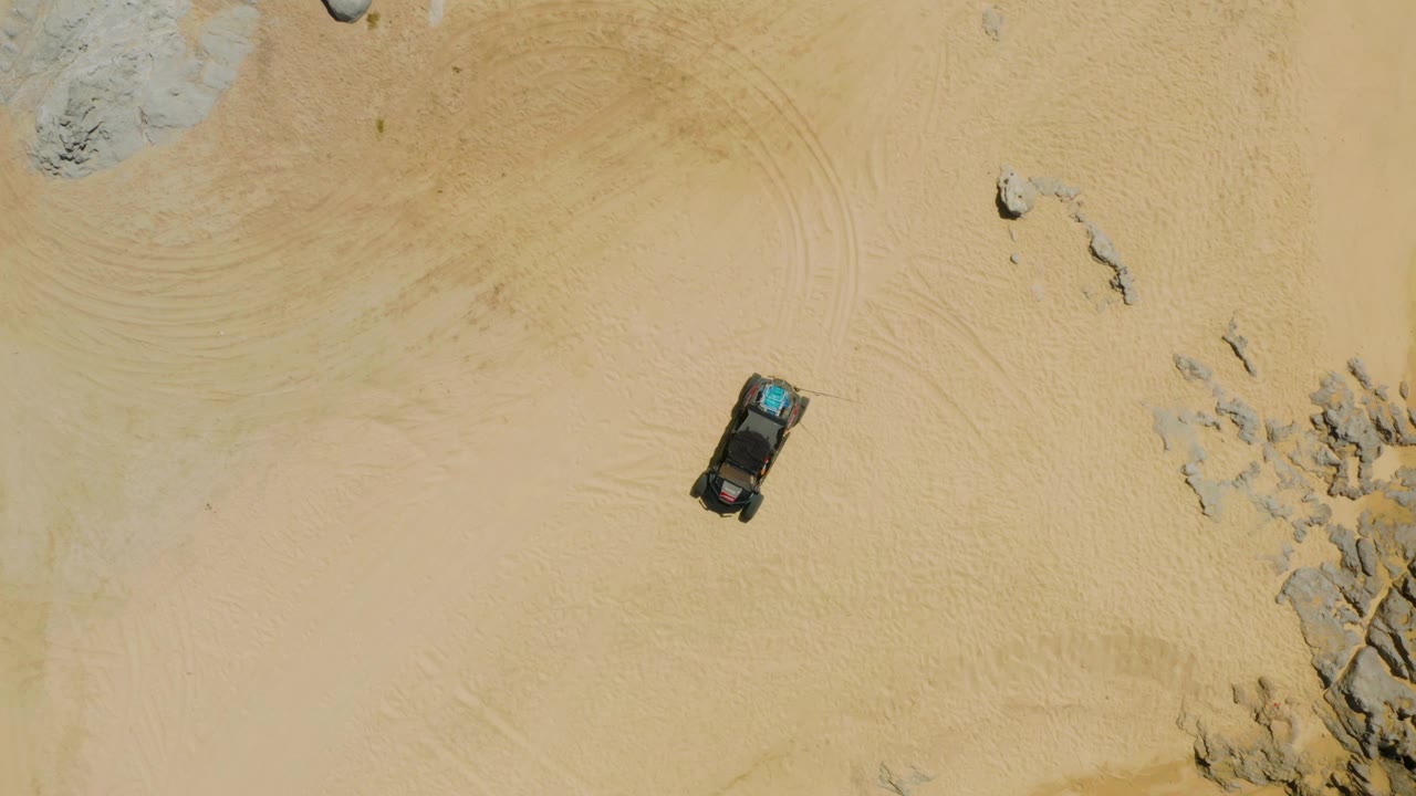 Aerial frame of the SUV slightly farther across the sand, with curved tire marks, scattered rocks, and no visible people.