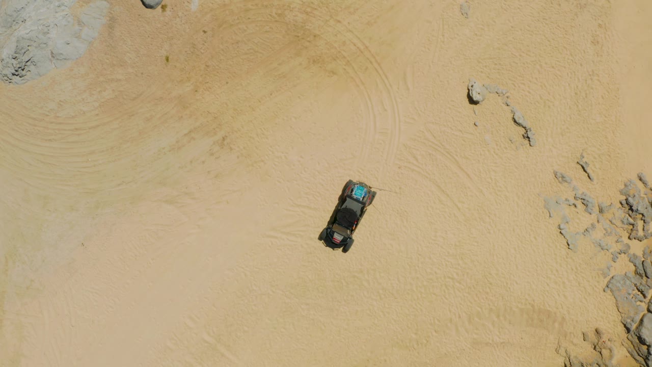 Top-down drone-like view of a dark SUV on tan sand in Laguna Beach, Baja California Sur, with rocks at the frame edge and no visible people.