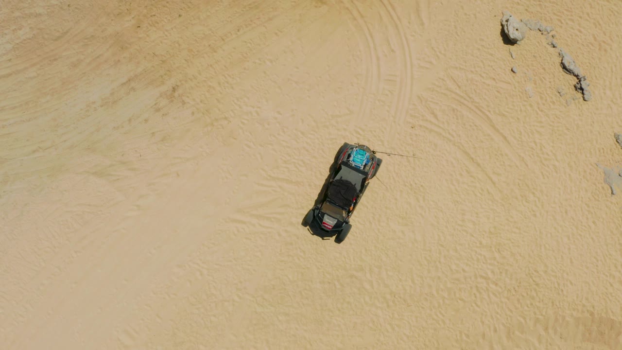 Overhead view of a dark vehicle parked on pale sand in Laguna Beach, Baja California Sur, with small rocks nearby and no visible people.