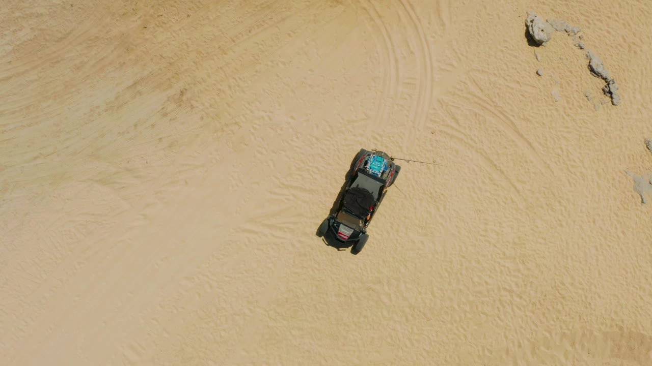 Aerial frame showing the parked dark vehicle below a small rock cluster on pale sand in Laguna Beach, Baja California Sur, with no visible people.