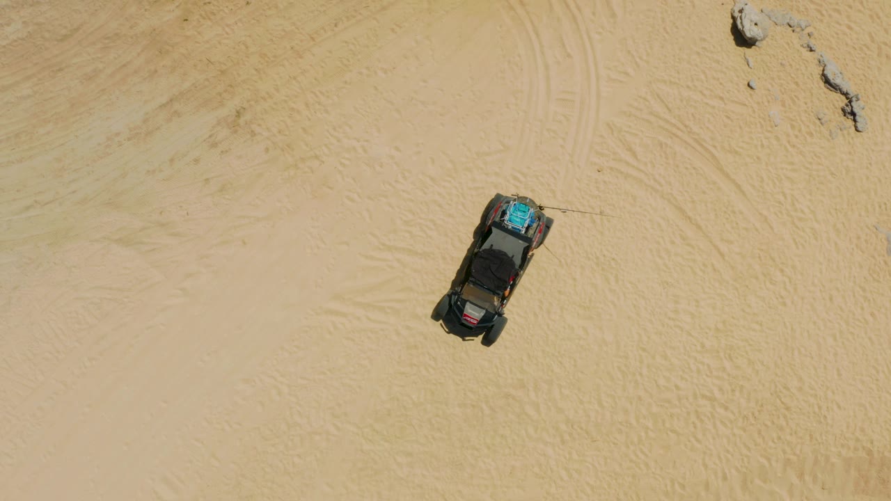 Steep downward drone view of a dark parked vehicle on open sand with a rock outcrop near the top of frame in Laguna Beach, Baja California Sur, and no visible people.
