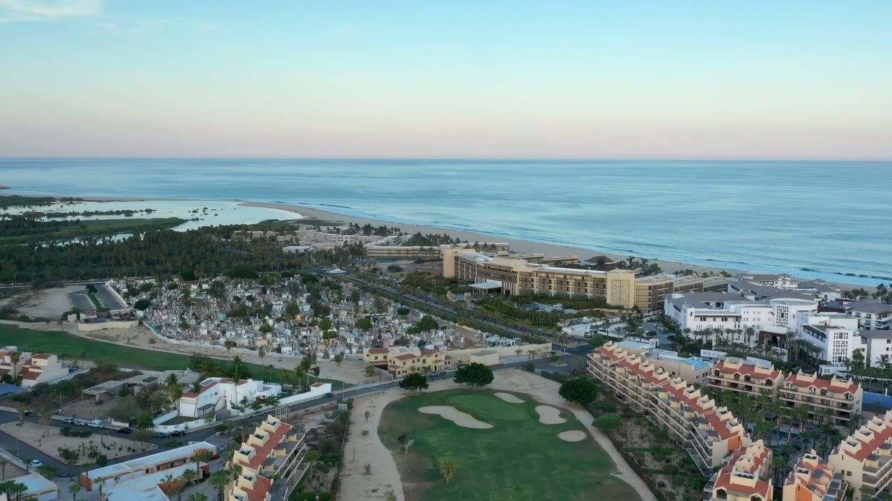 A second aerial view of San Jose del Cabo with the ocean horizon high in frame, resort-style buildings near the beach, homes below, golf fairway, and no visible people.