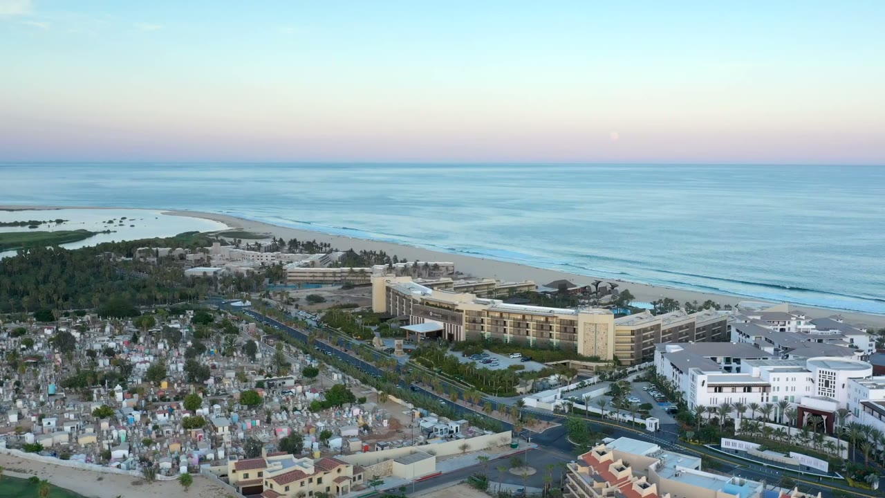 Aerial view of coastal buildings, beach, ocean, and foreground cemetery-like markers.