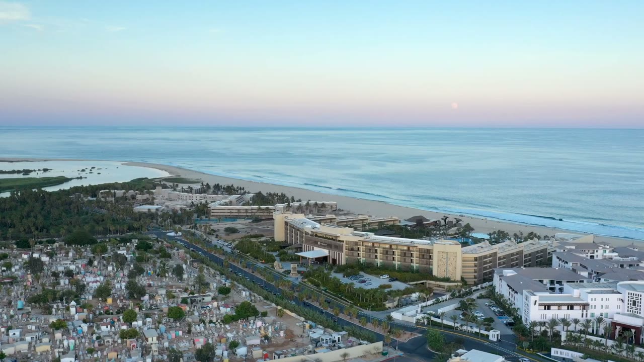 Aerial frame of San Jose Del Cabo showing the beach and ocean behind large coastal buildings, with roads and cemetery-like markers below and no visible people.