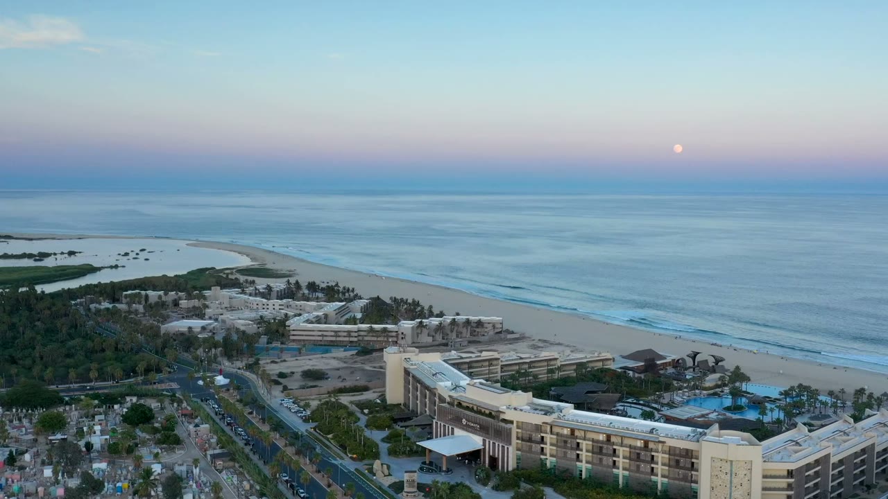 Elevated dusk view of beach, ocean waves, resort buildings, and a small moon.