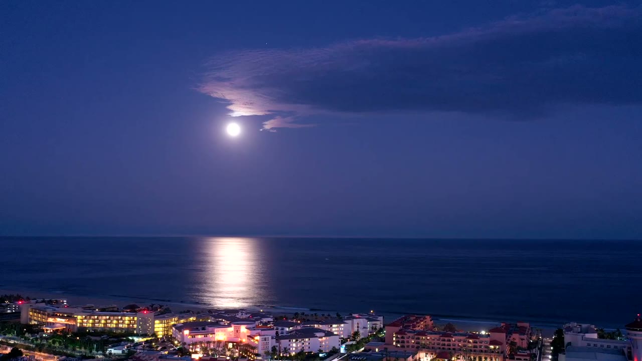San Jose del Cabo night coastline with a bright moon partly veiled by cloud, a vertical reflection on the ocean, lit buildings below, and no visible people.