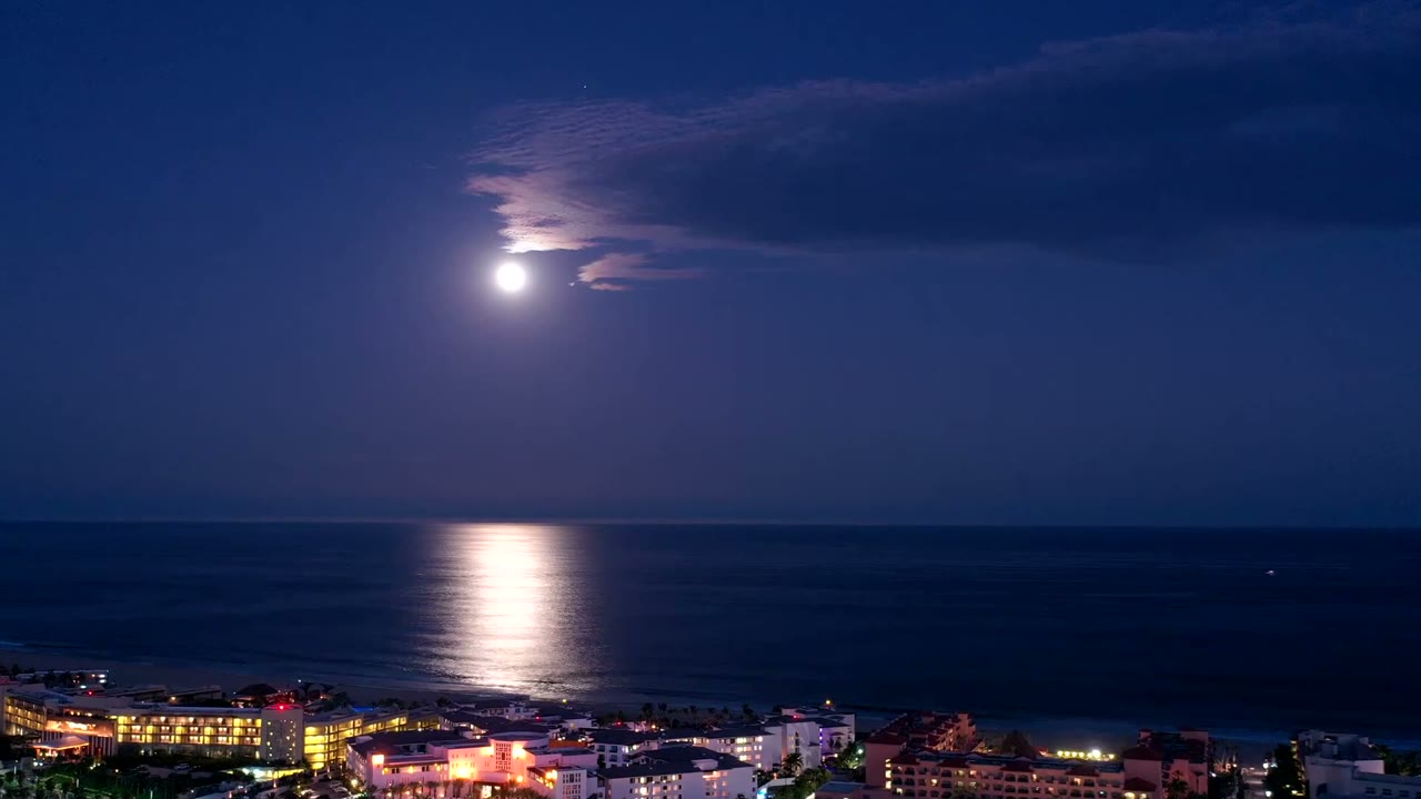 San Jose del Cabo coast at night with the moon near a cloud bank, dark ocean across the center, warm city lights along the bottom, and no visible people.