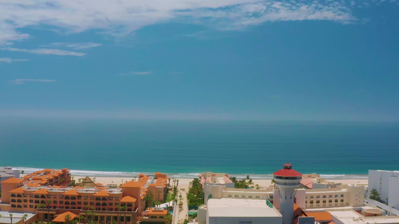 Elevated Palmilla Beach frame with the ocean horizon high in the background, surf along the sand, clustered orange roofs, white hotel structures, palms, and no visible people.